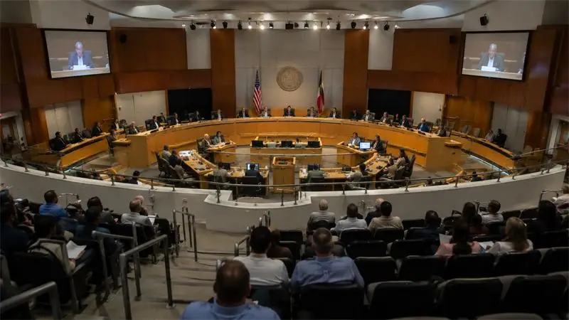 Councillors sit in a circular chamber during a full council session, watched from the public gallery.